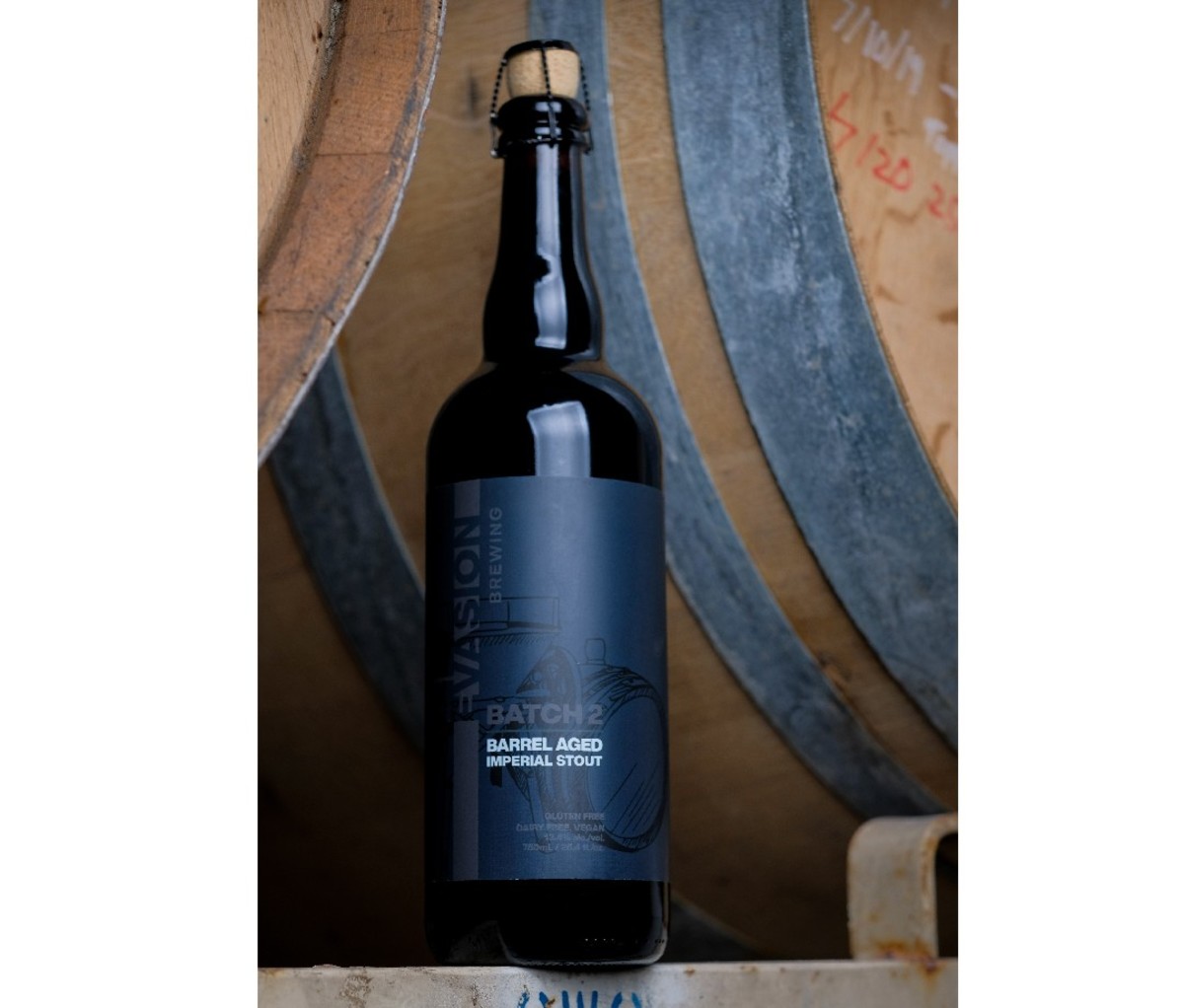 Tall black bottle of Evasion Brewing Batch 2 Barrel Aged Imperial Stout in front of a barrel wall background.
