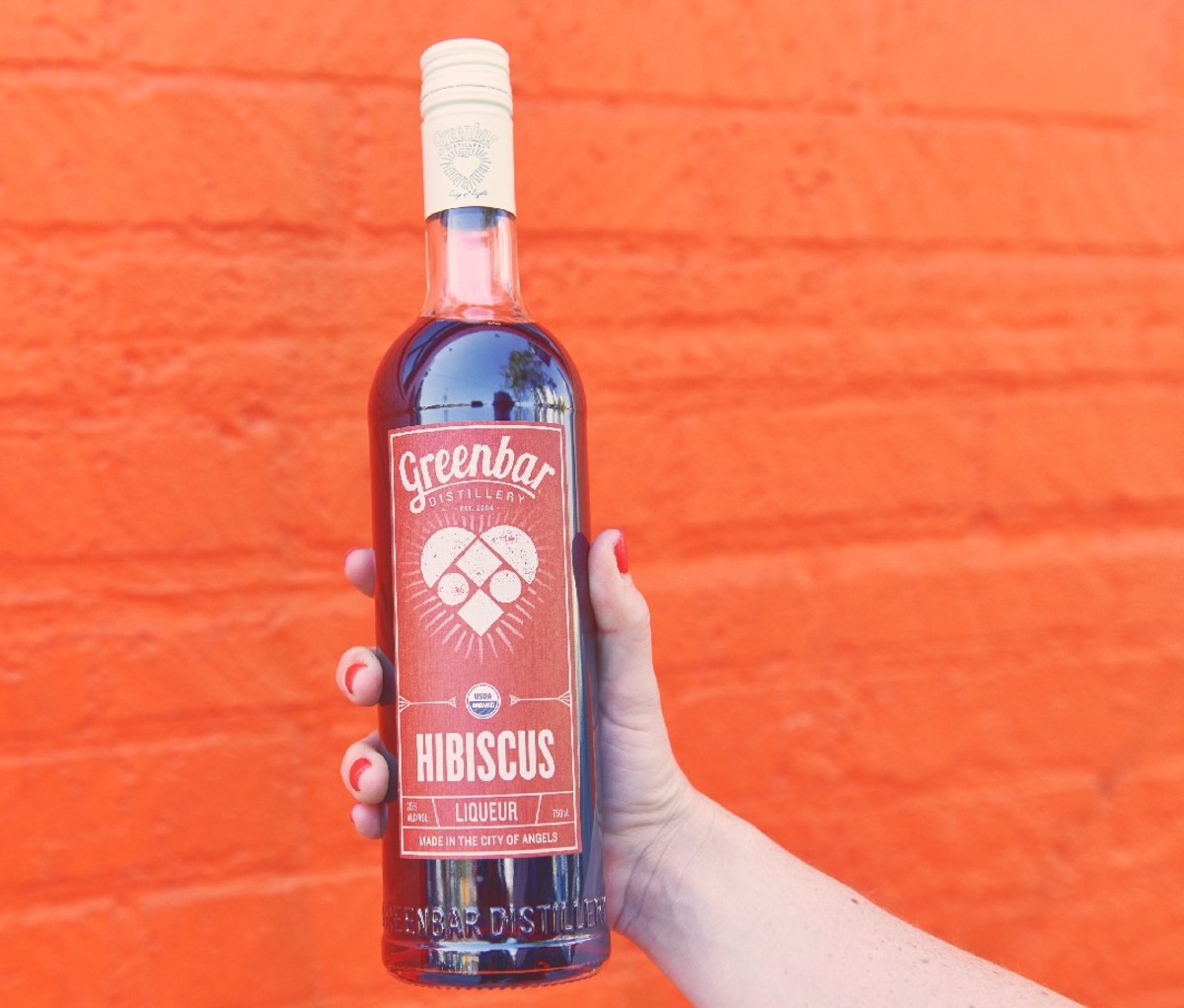 Bottle of Hibiscus Liqueur held in an outstretched hand in front of an orange wall