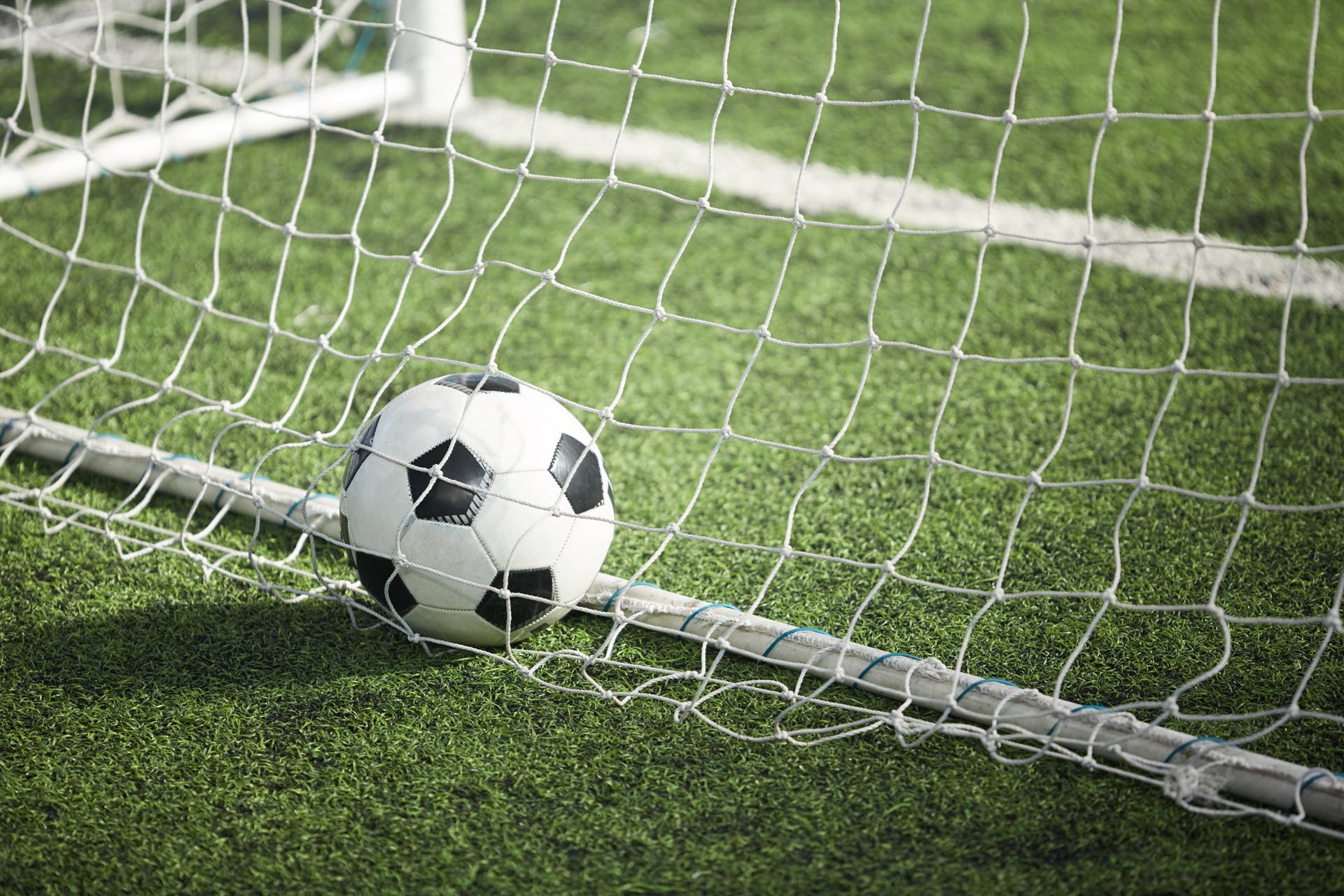 Leather soccer ball in empty goal behind a net on green soccer field.