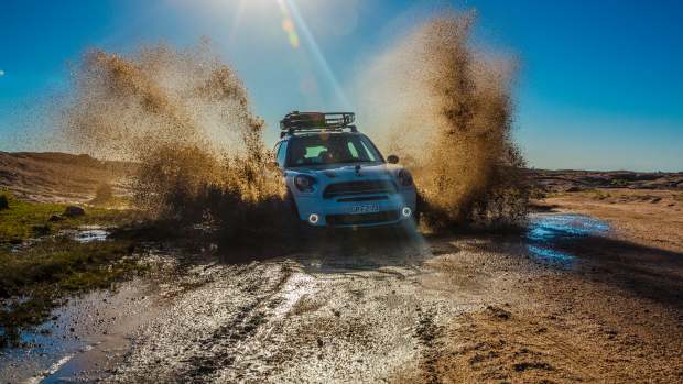Small AWD Mini car driving off-road through mud spraying it into the air with a blue sky and sun in the background.