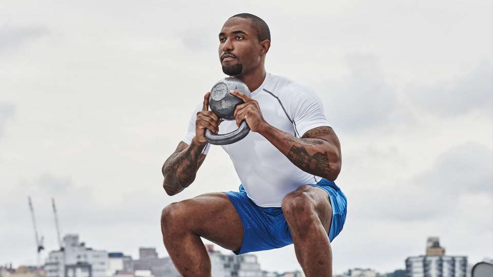 Black man wearing white T-shirt and blue shorts on urban rooftop doing goblet squat