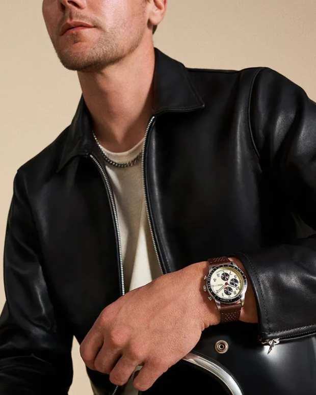 Man in a black leather jacket wearing a chronograph watch on a tan background with two other watches next to him on a white to brown gradient background.