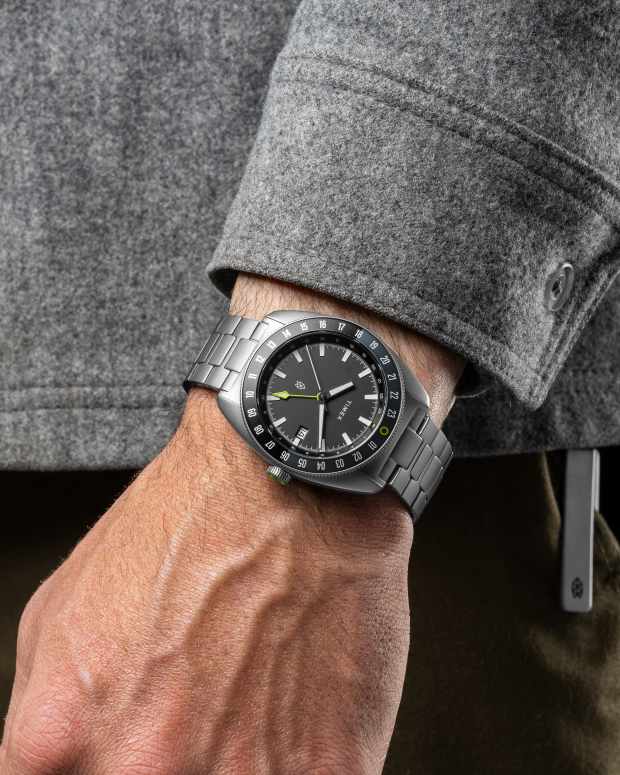 Man wearing a gray titanium watch from James Brand and Timex with a gray long-sleeve shirt on and green pants on a black background, watch news from Men's Journal.