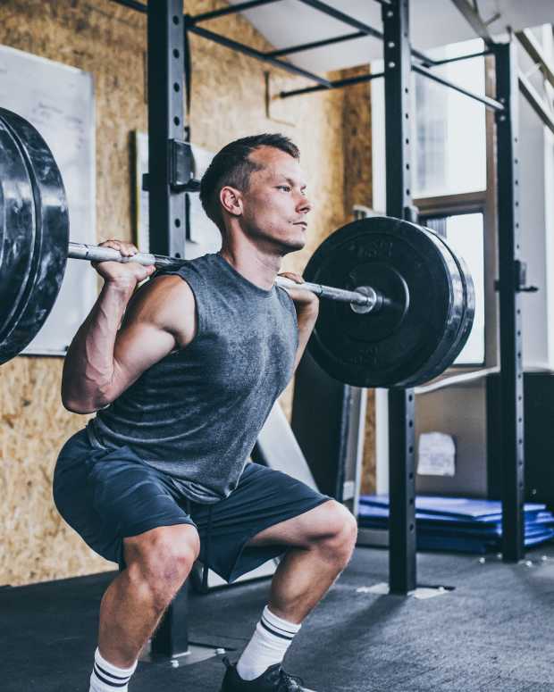 Man in gray tank and navy shorts performs barbell back squat in the gym.