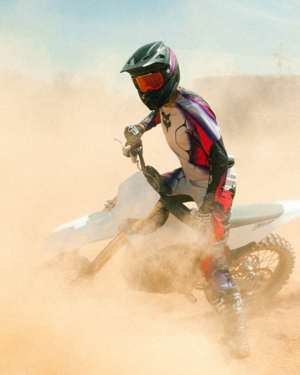 Dirt bike rider in black and red outfit and helmet on a white Stark VARG e-motorcycle doing donuts on sandy ground, new motorcycle review.