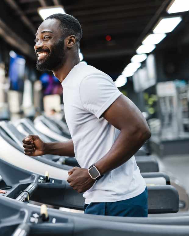 Man with white t-shirt and blue shorts walks on a treadmill with a smartwatch.