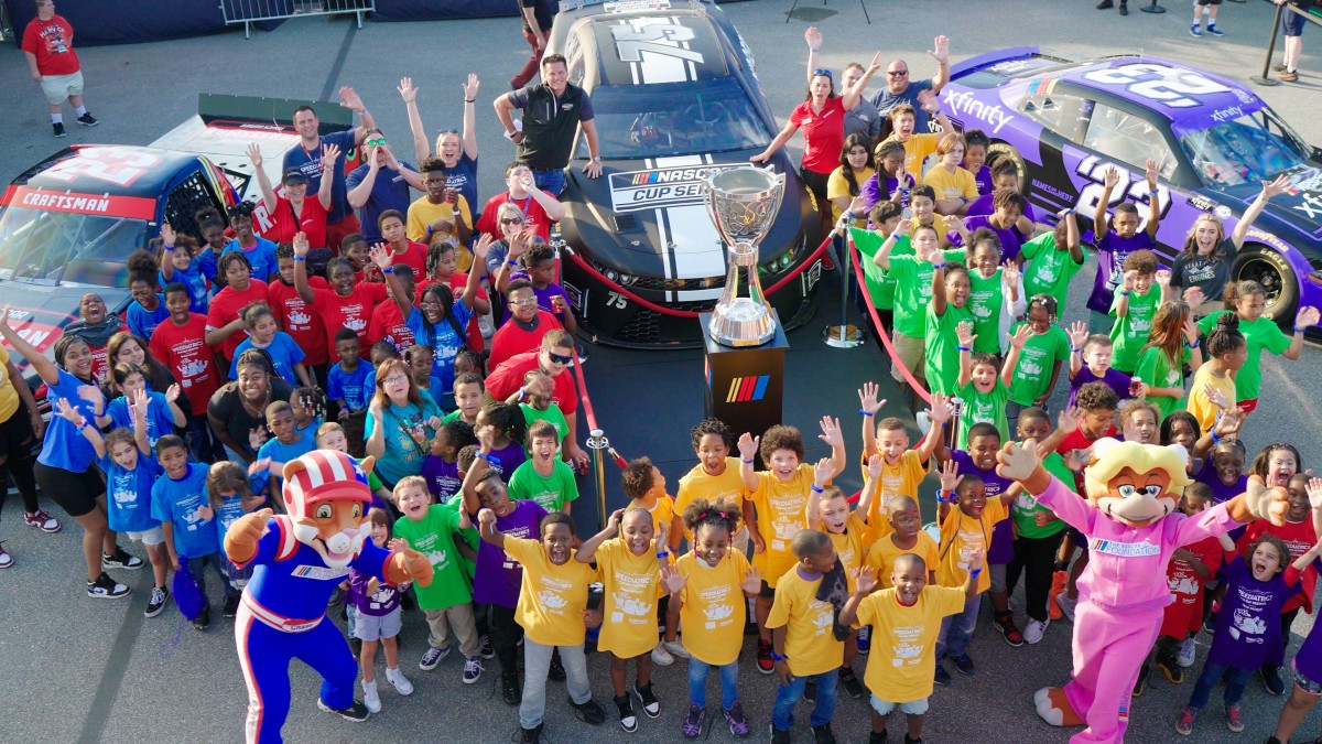 Kids in groups wearing red, blue, yellow, and green T-shirts pose excitedly in front of NASCAR cars