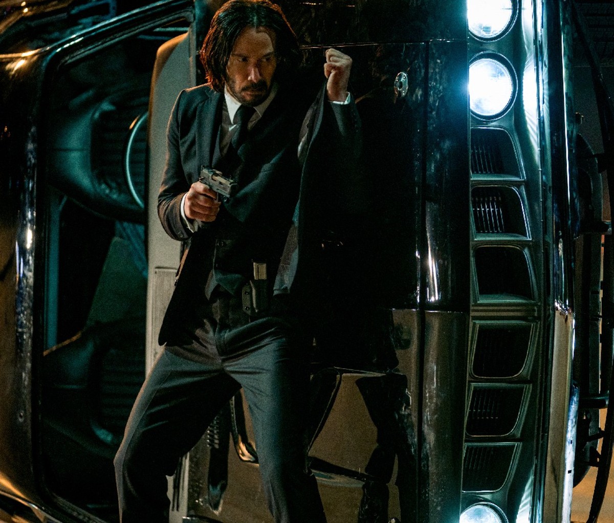 Keanu Reeves takes cover behind a car with gun in hand in a scene from John Wick: Chapter 4