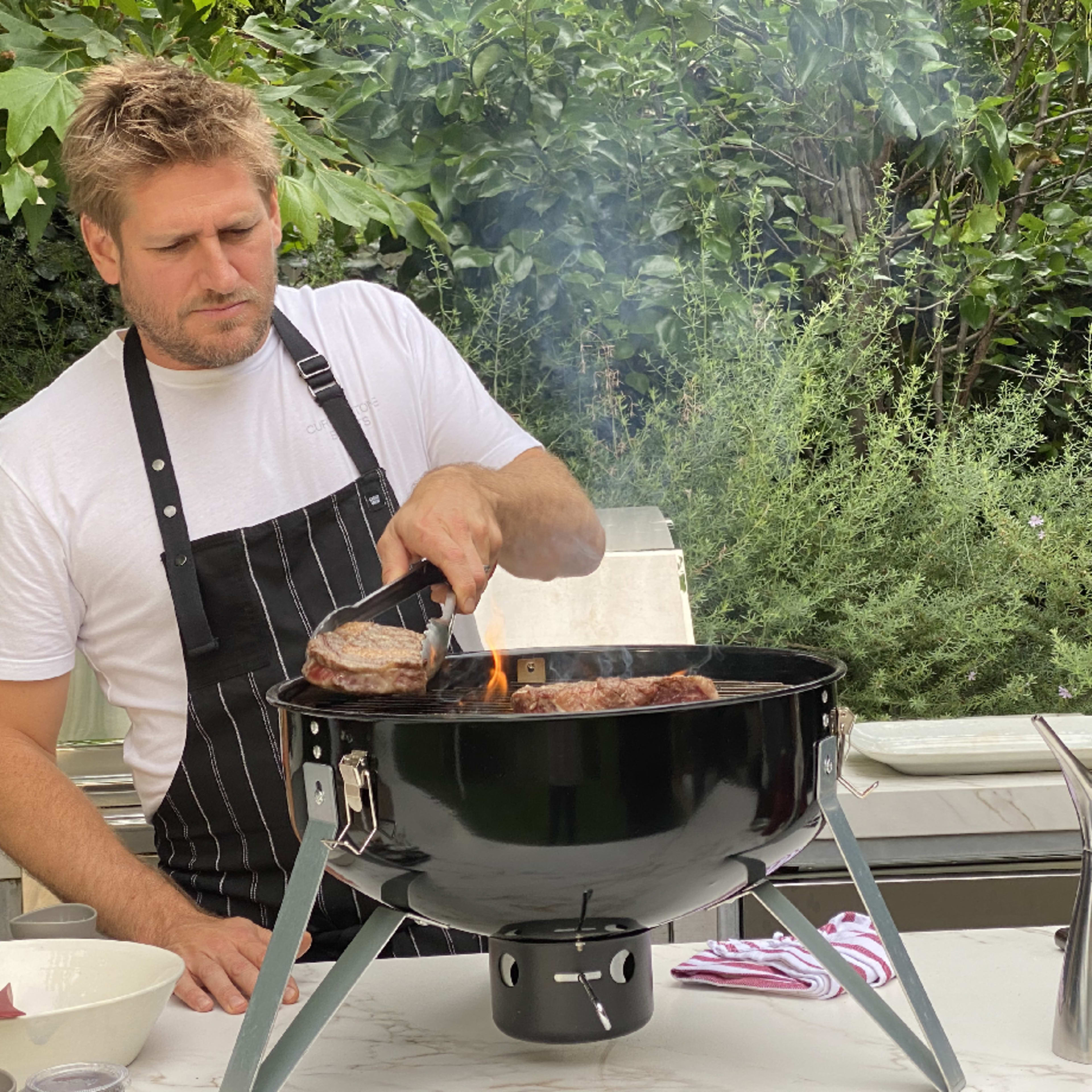 Chef Curtis Stone using tongs to move a piece of steak cooking on a small kettle grill with bushes in the background, part of our 2024 Men's Journal grilling guide and awards.