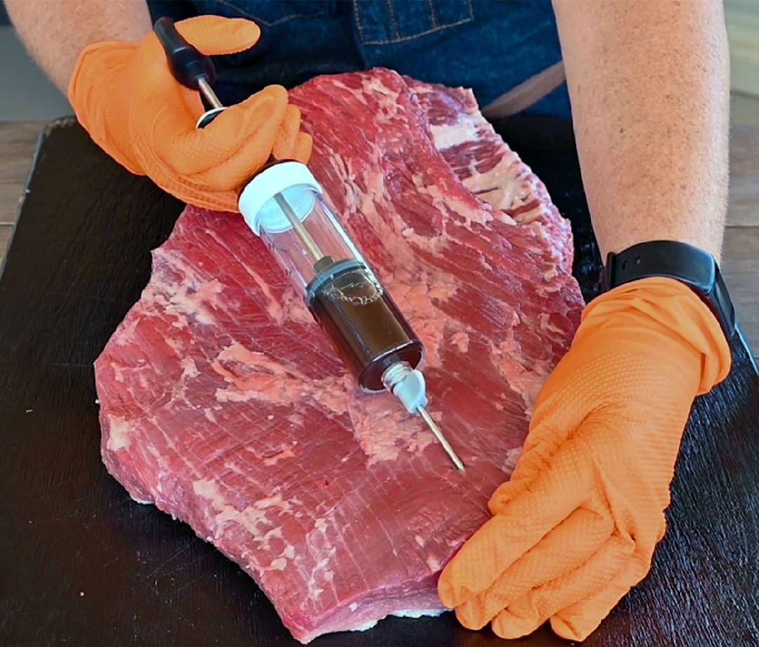 Person with orange gloves on injecting beef broth into a beef brisket.
