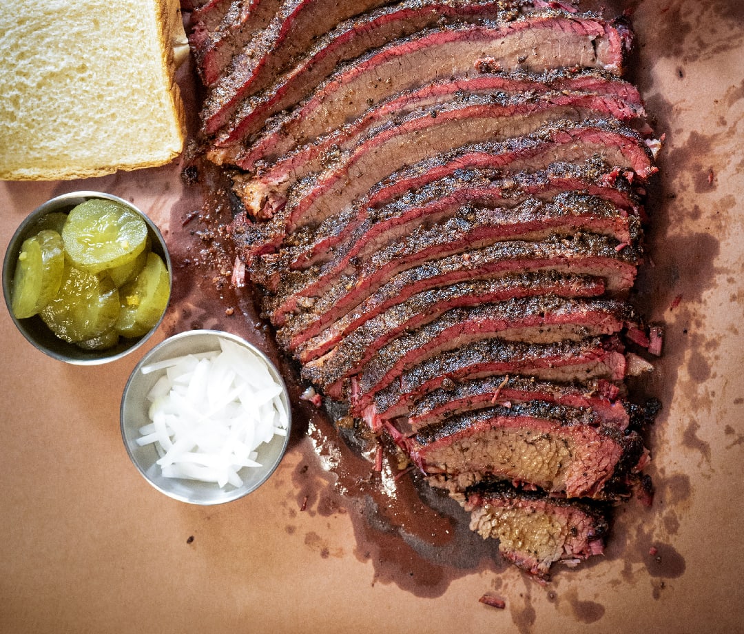 Slices of brisket on a brown butcher paper with white bread, pickles and onions.