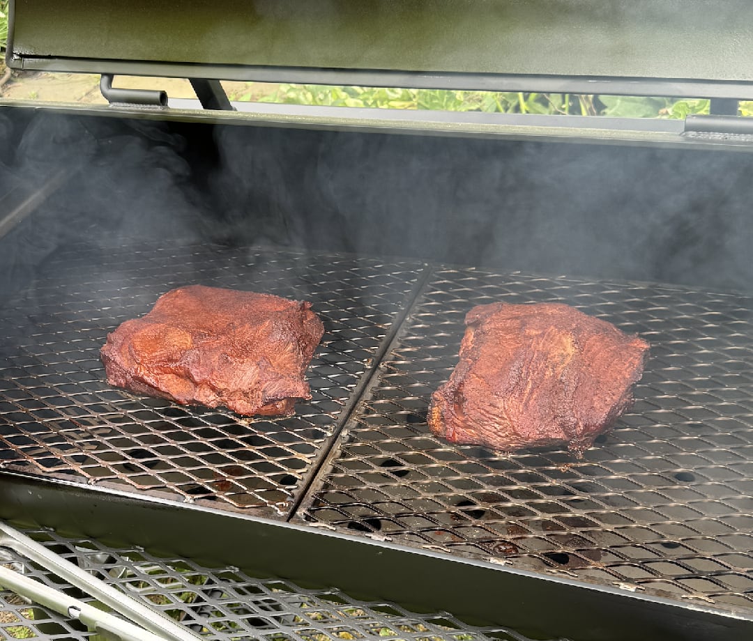 Two beef briskets smoking on a smoker grate with smoke.
