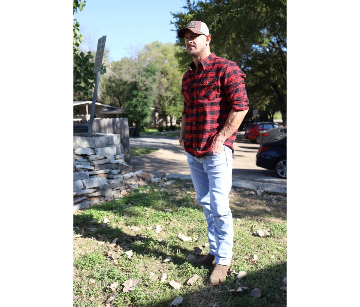 Man in red flannel shirt, light blue jeans, suede boots, and baseball hat standing on grass