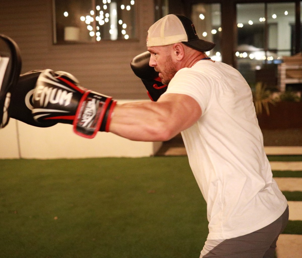 Caucasian man in white T-shirt and backwards baseball cap punching punching bag with boxing gloves