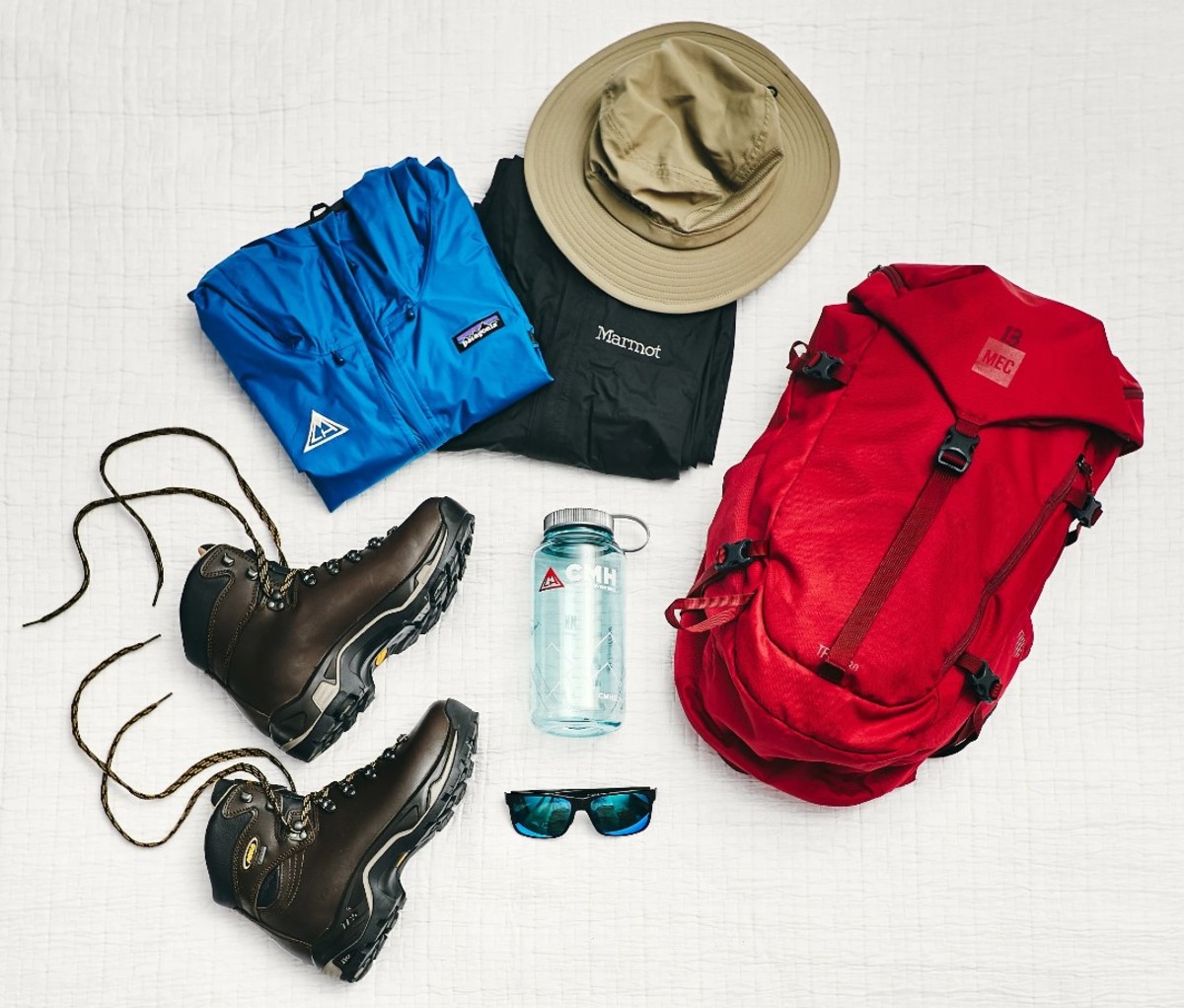 Hiking boots, blue and black jackets, sun hat, red backpack, sunglasses, and water bottle on white backdrop