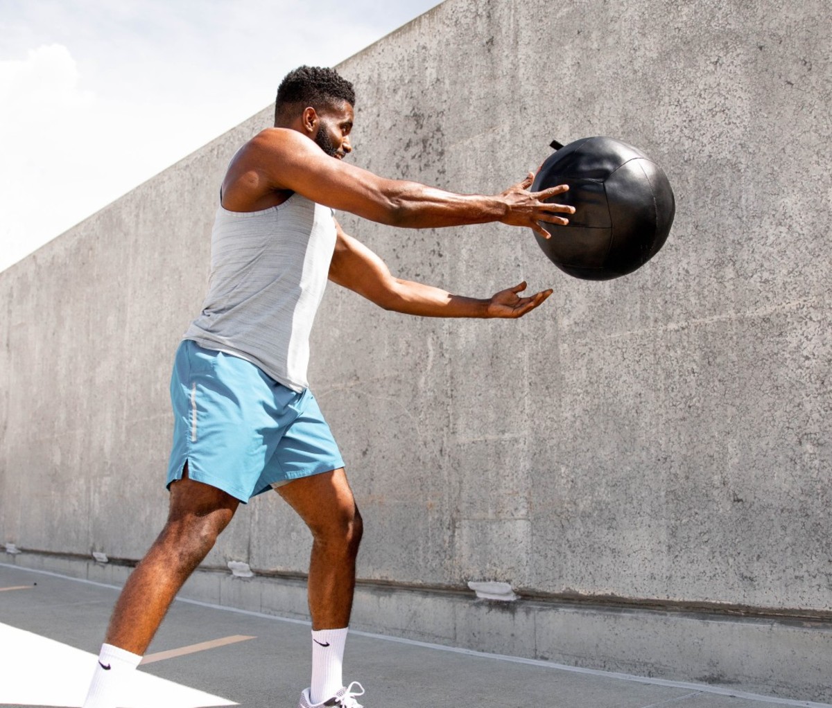 Black man wearing white tank top and blue shorts throwing medicine ball against wall