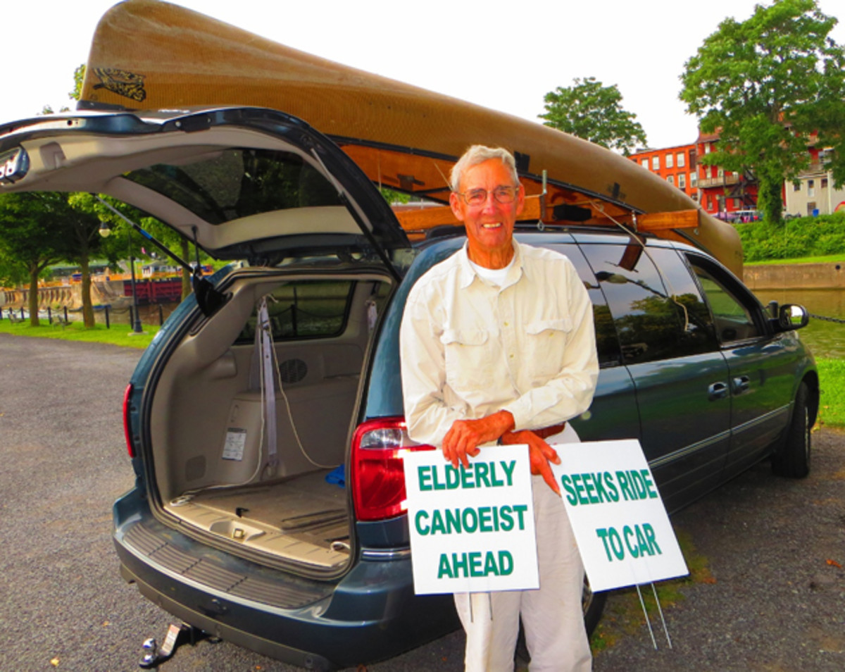 John Stookey stands before his Souris River Quetico while showing of his hitch hiking signs. Photo courtesy of Robert Stopper