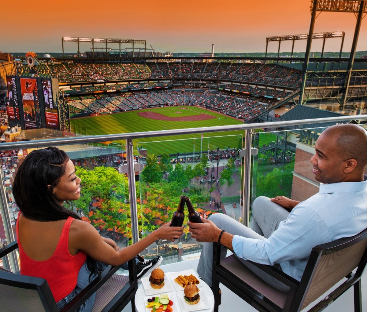 Black couple cheersing beers on balcony overlooking baseball stadium