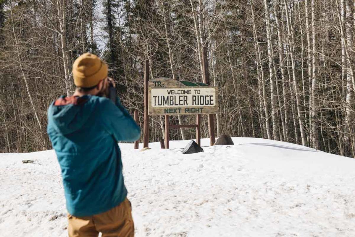 A man in a blue jacket and yellow hat takes a photograph of the Tumbler Ridge welcome sign in the snow.