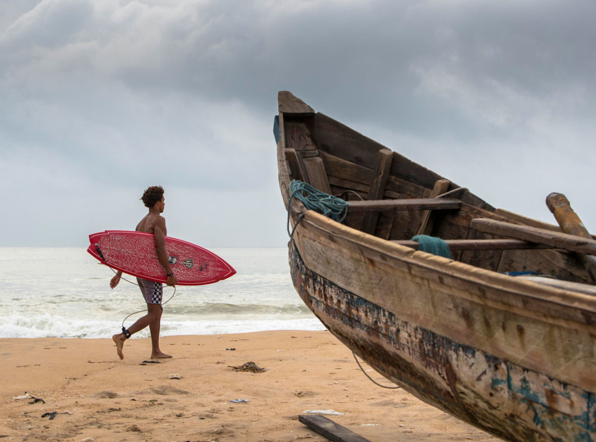 Michael February walking with surf board on beach in Ghana