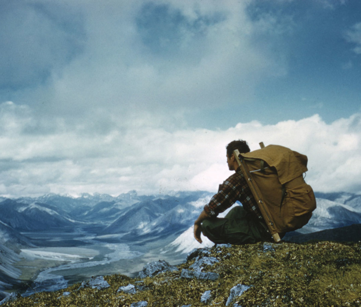 Backpacker sitting cross-legged on edge of cliff overlooking Alaskan mountain range