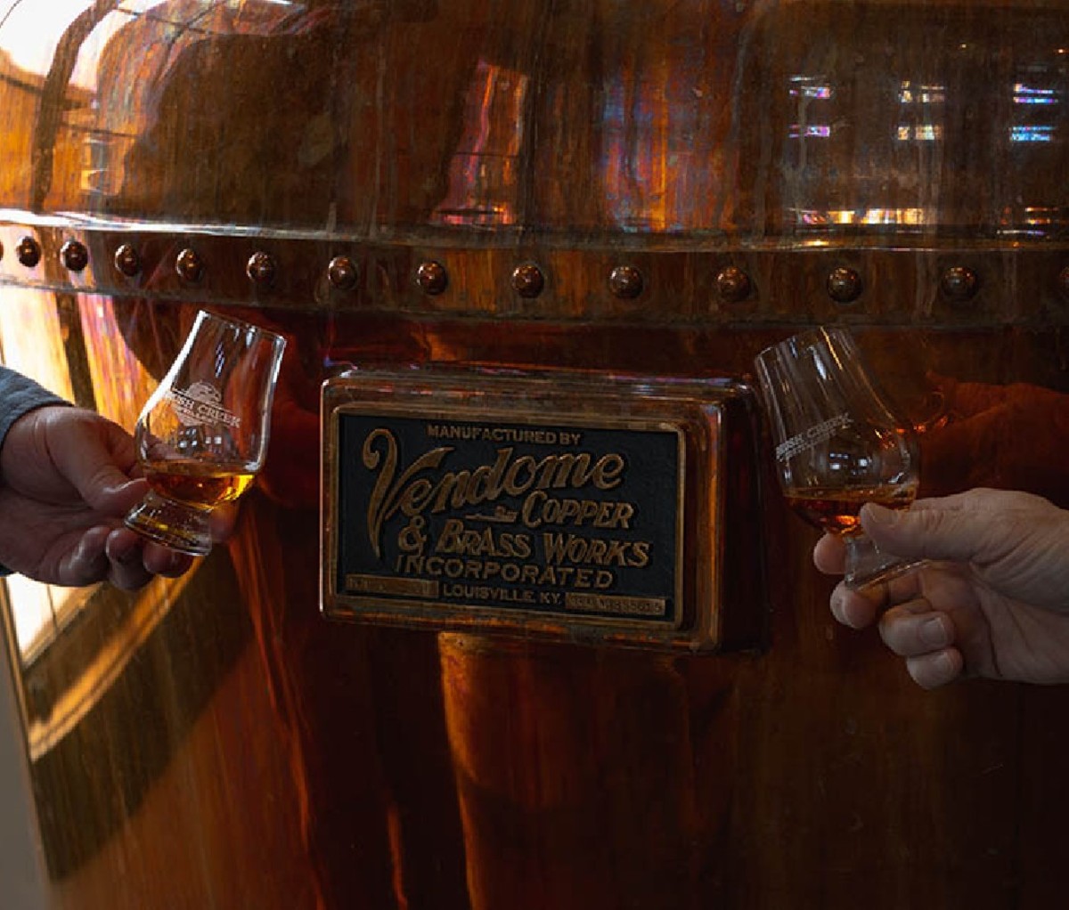 Two hands holding Brush Creek whiskey in front of a large copper still.