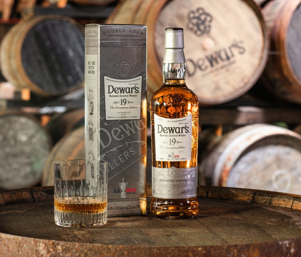 In front of whiskey casks, a box, bottle and glass full of Dewar’s 19-Year-Old The Champion’s Edition.