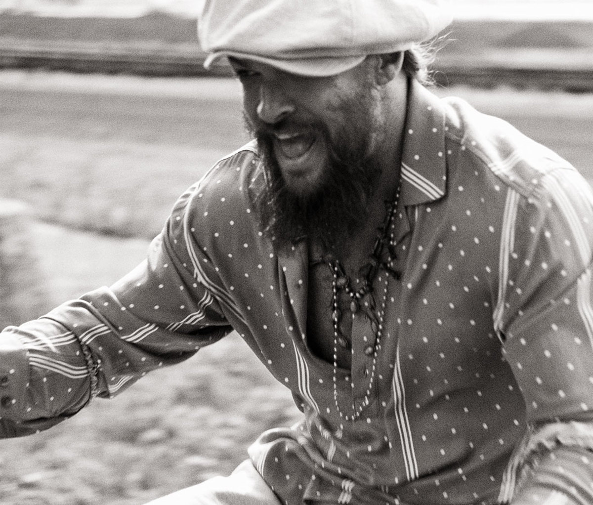Black and white photo of Jason Momoa riding bike wearing cabbie hat