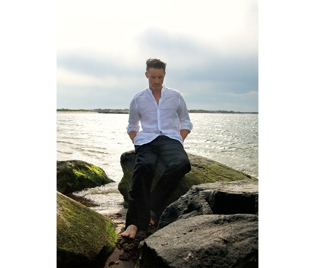 Man in white linen button-down shirt sitting on rocks by beach