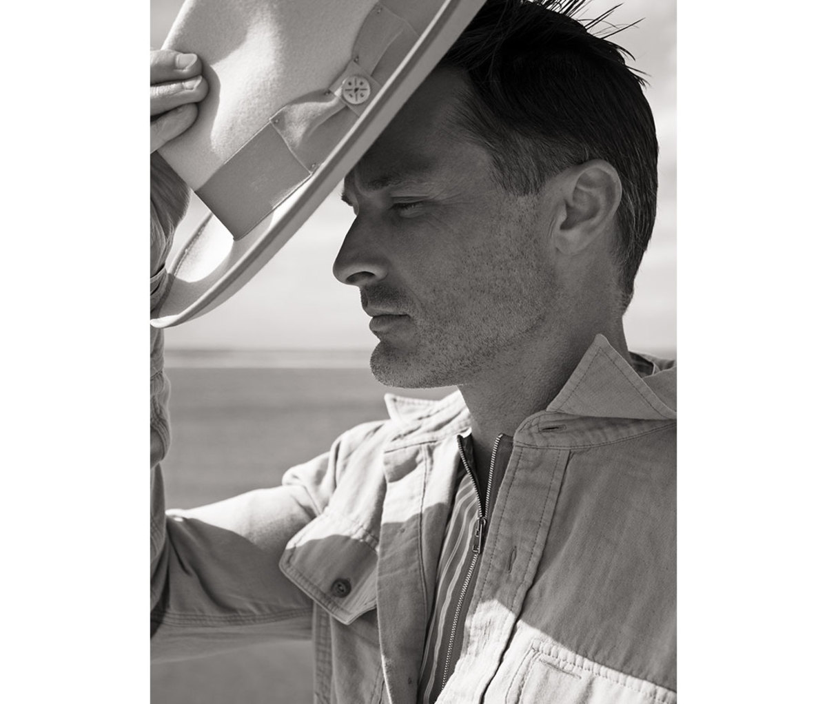 Black and white portrait of man putting hat on with ocean in background