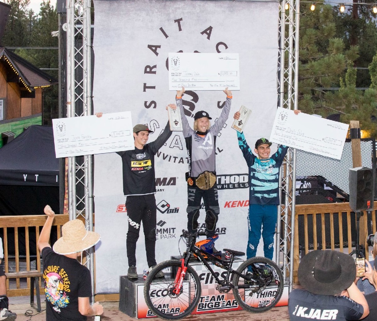 3 bike racers on the winner's podium holding giant checks above their heads.