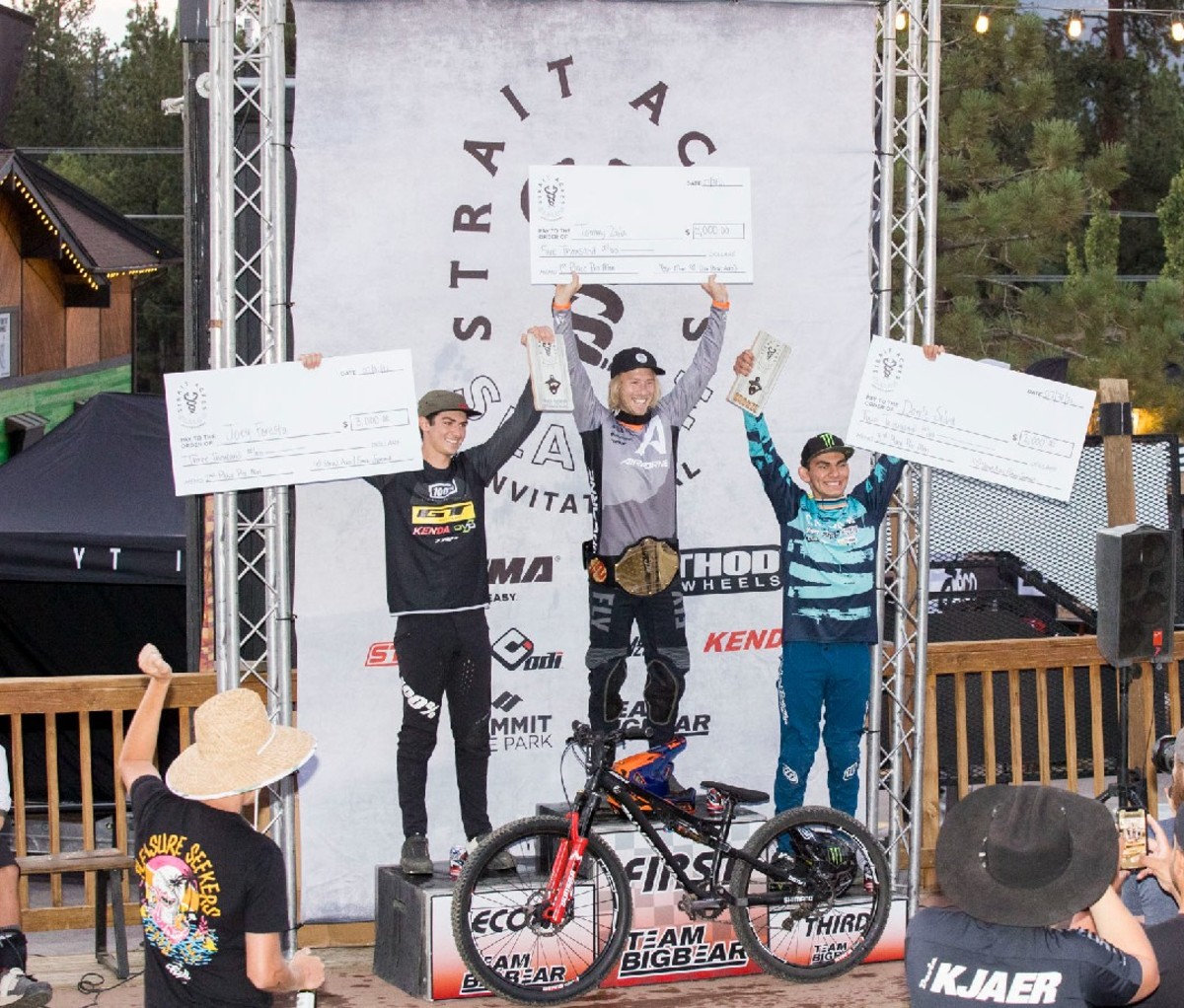 3 bike riders on the winning podium with giant checks over their heads.