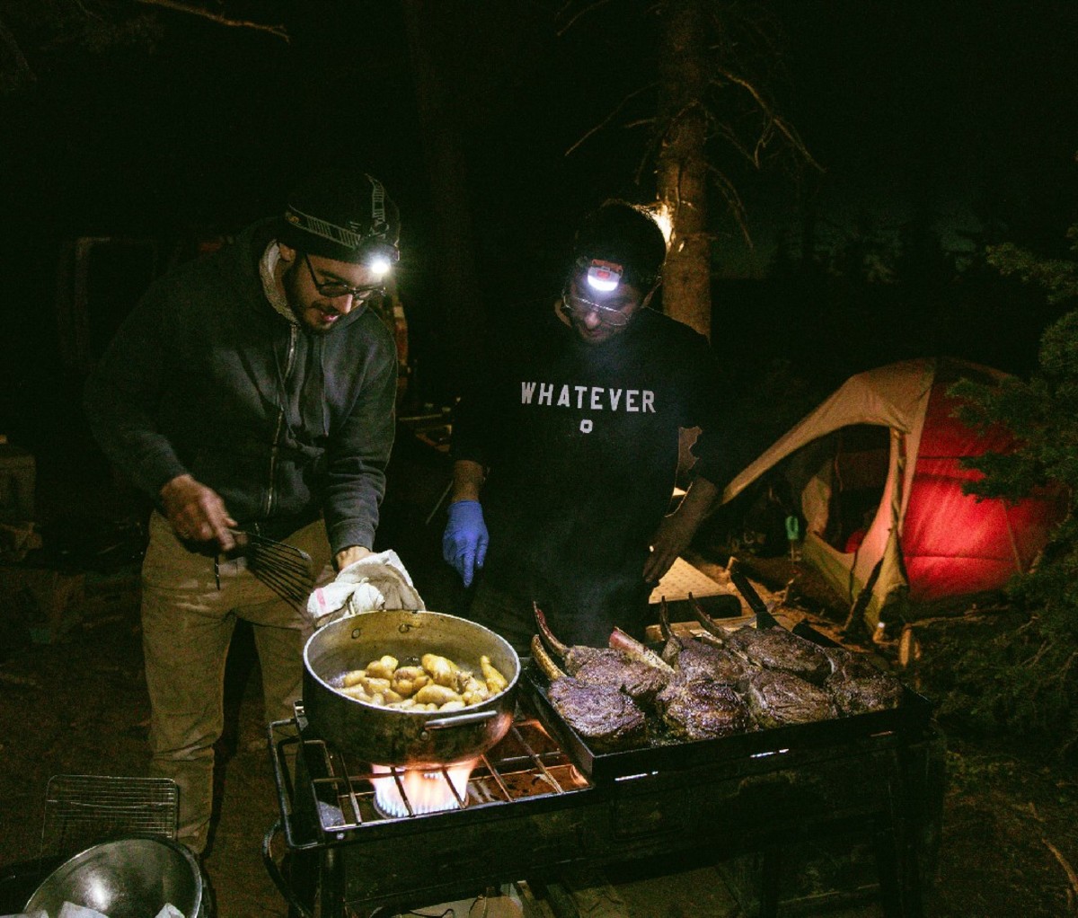 Two men cooking potatoes and steaks with headlamps on in campsite