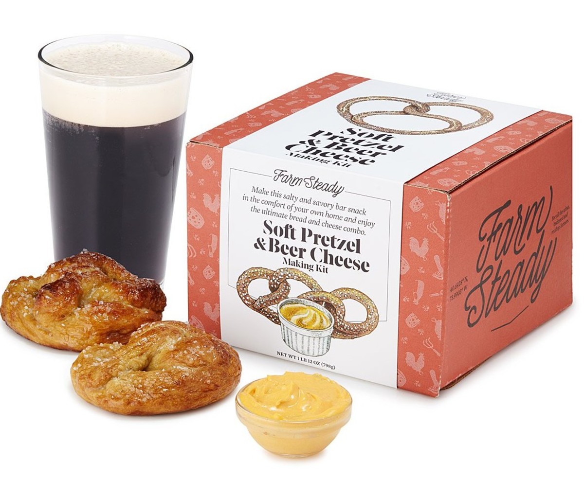 Uncommon Experience's Soft Pretzel & Beer Cheese Making Kit in a box, displayed beside two pretzels, a small bowl of soft cheese and a pint of beer