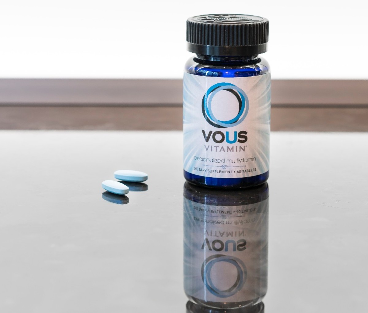 Vous Vitamin bottle on a glass table beside two light blue vitamin pills/supplements
