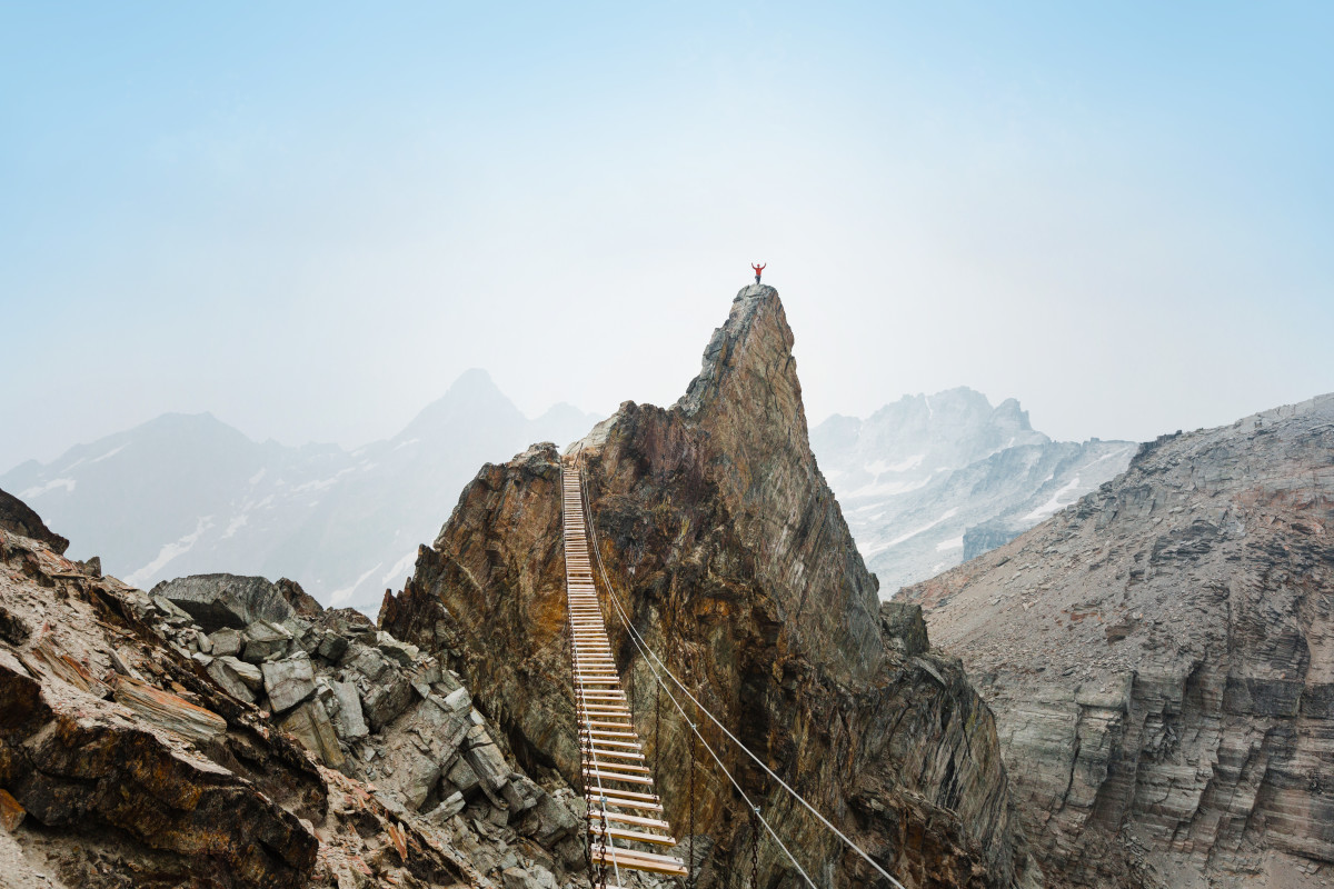 Hiker in distance stands on a mountaintop with arms raised on the far from a slat bridge across a precipice on the Mt. Nimbus Via Ferrata in the Western Canadian mountains