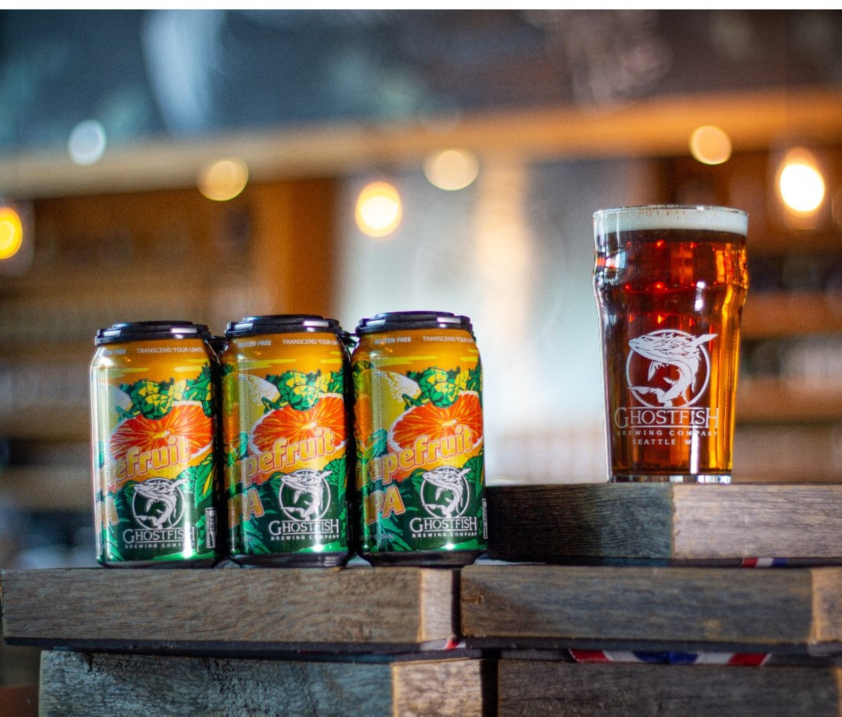 Three cans of Ghostfish Brewing Company Grapefruit IPA on a wooden table next to a filled pint glass.