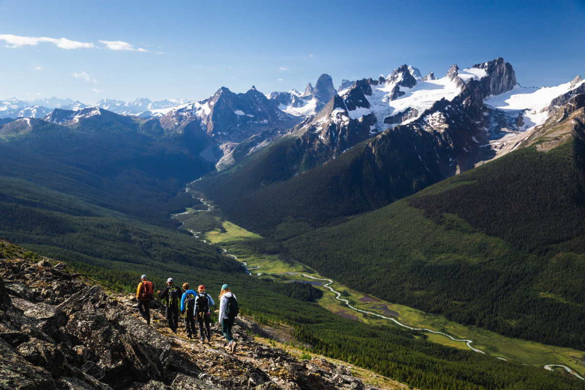 A group of five hikers walk along a mountain trail above a green valley with snowy peaks in the background