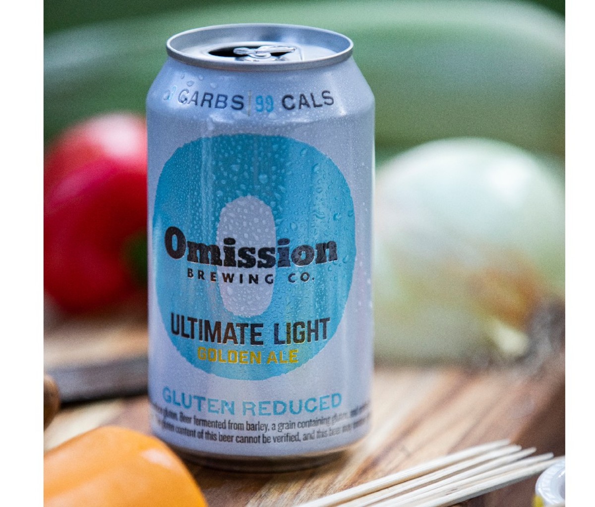 Can of gluten-reduced Omission Balanced Brewing Ultimate Light Golden Ale on a cutting board with vegetables in the background.