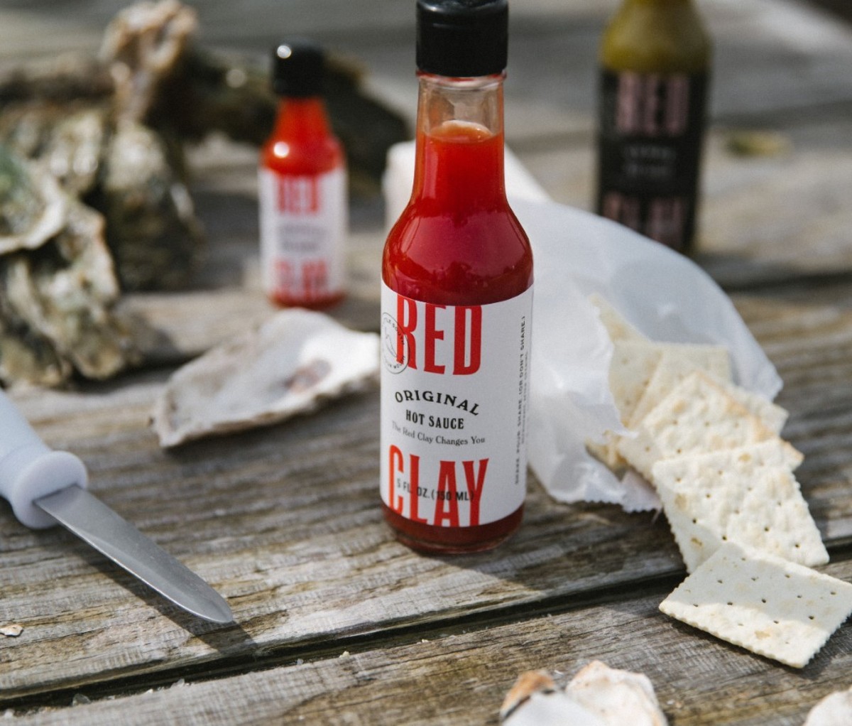 Red Clay Original Hot Sauce surrounded by a pack of crackers and a knife on a wooden table.