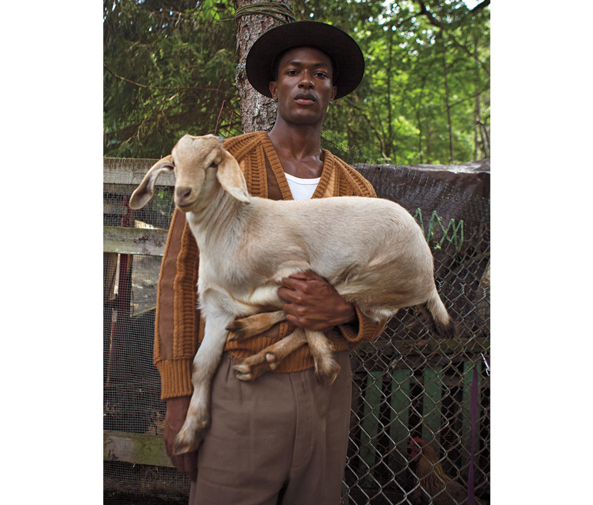Black man in hat and auburn sweater holding a goat