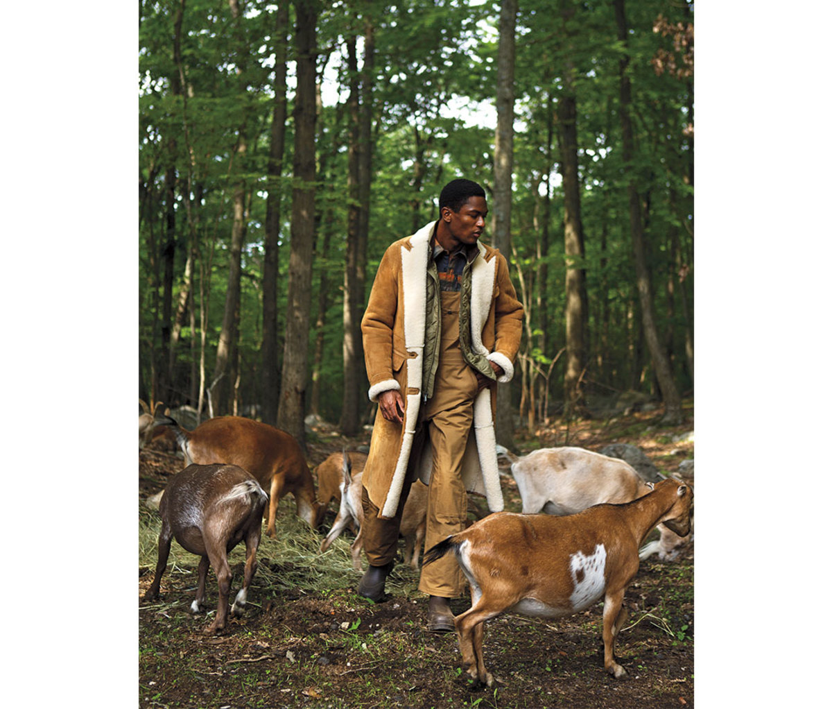 Black man wearing shearling coat in forest with goats