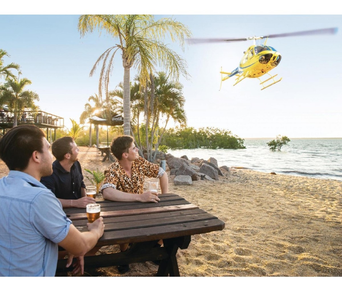 Three men at a table drinking beer watch a yellow helicopter landing on the beach in Northern Australia