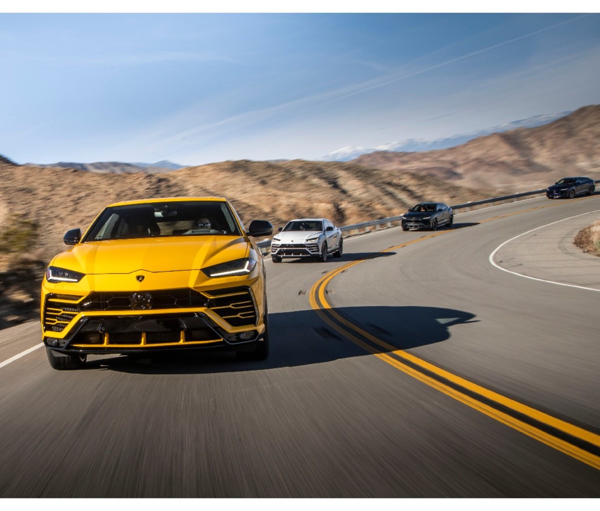 Front of yellow 2021 Lamborghini Urus driving on a curved desert road with three Lambo Urus's behind