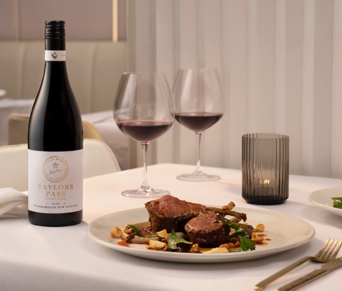 Bottle of 2018 Villa Maria Single Vineyard Taylors Pass Pinot Noir on a white linen table next to a dinner plate and two glasses of wine