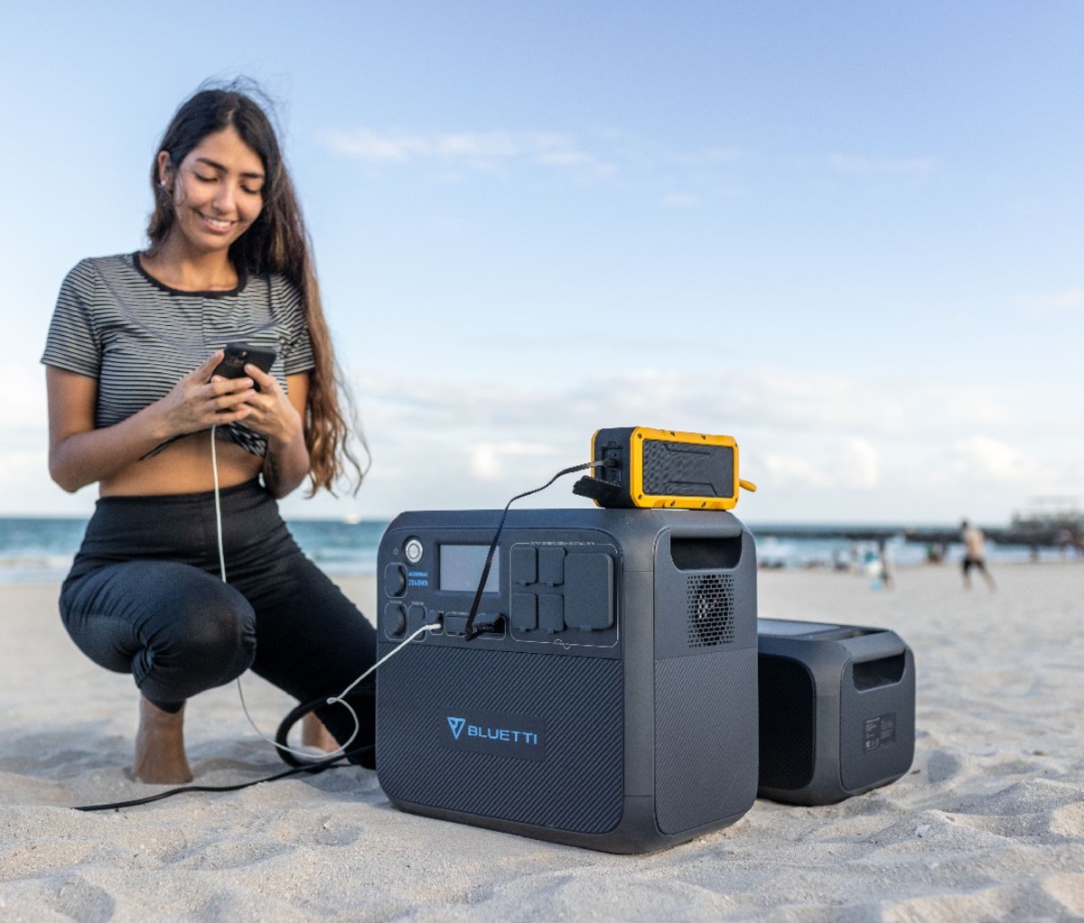 Woman charging her phone on the beach beside Bluetti portable power station