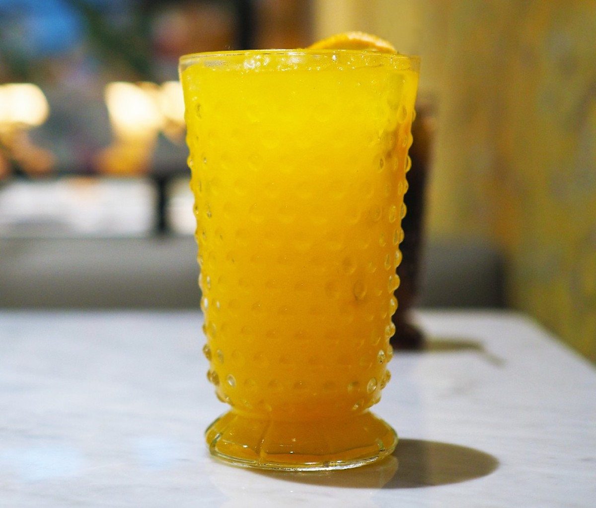 Bright orange-colored yuzu spritz in a tall glass with an yuzu garnish, on a gray slate surface