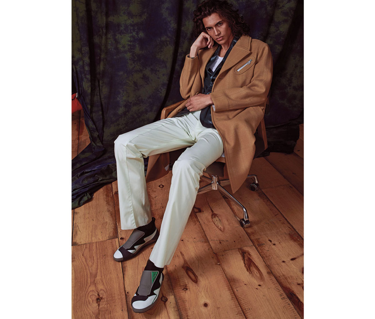 Man with shoulder-length hair sitting in chair wearing camel-colored coat, white pants, and sneakers