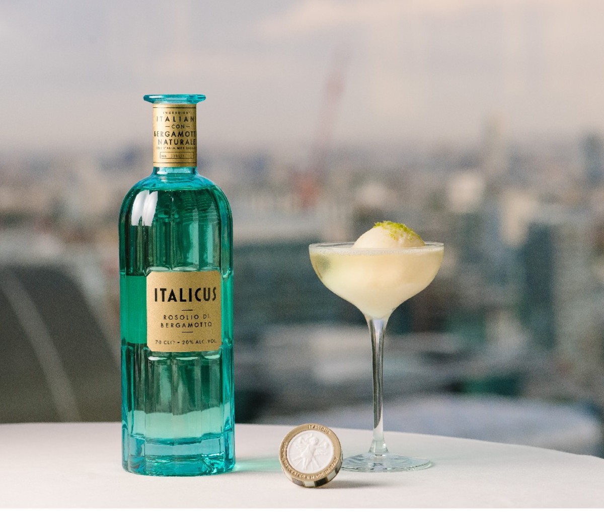 Bottle of Italicus Rosolio di Bergamotto on a table beside a cocktail in a coupe glass