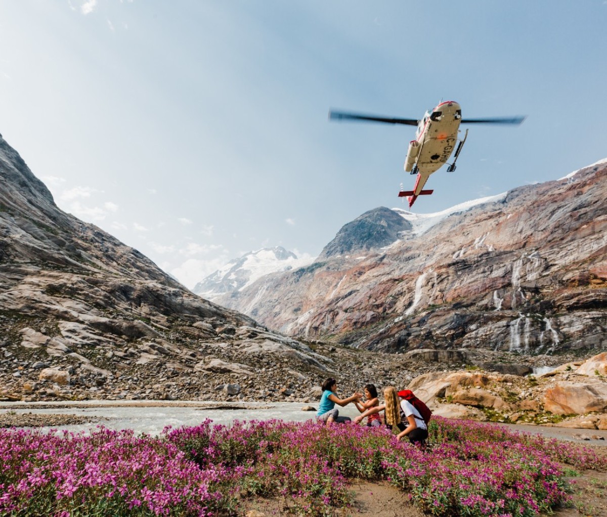 Helicopter flies over hikers at a remote mountain lake with pink wildflowers